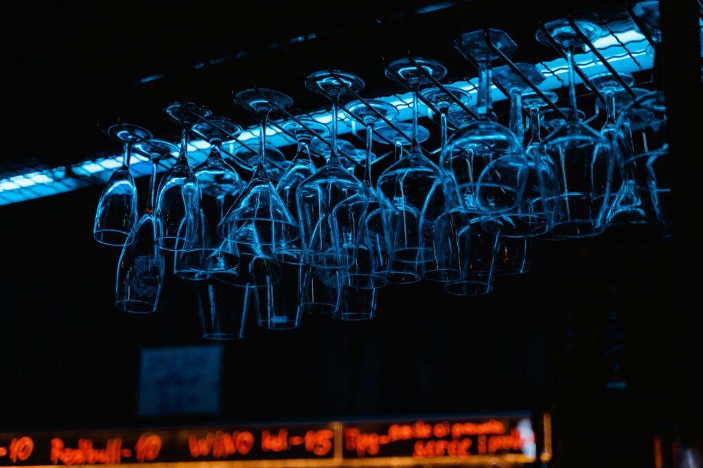 Racks of wine glasses hang in a dimly lit bar, illuminated by neon lights.